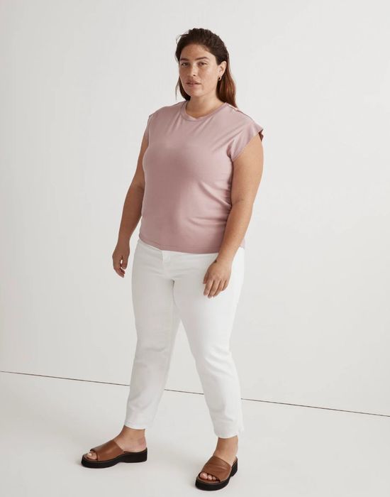 A person stands confidently in casual attire, sporting a sleeveless top, white pants, and sandals, in a minimalist environment