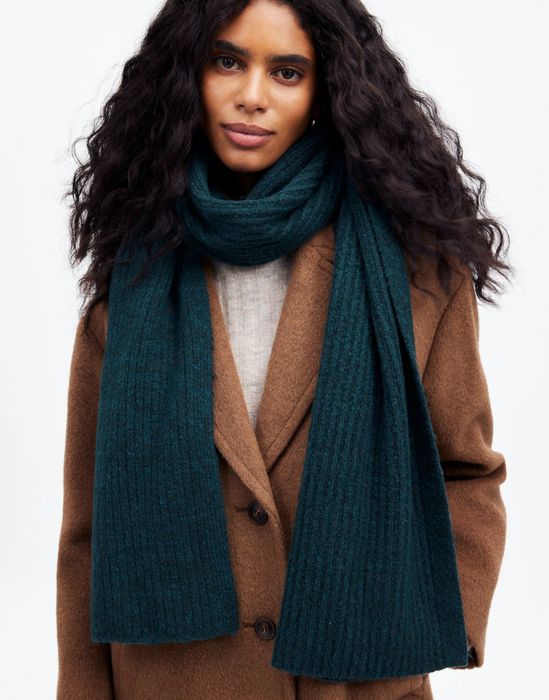 A person dons a brown coat and a long, thick scarf. Their long, wavy hair frames their face as they look directly at the camera