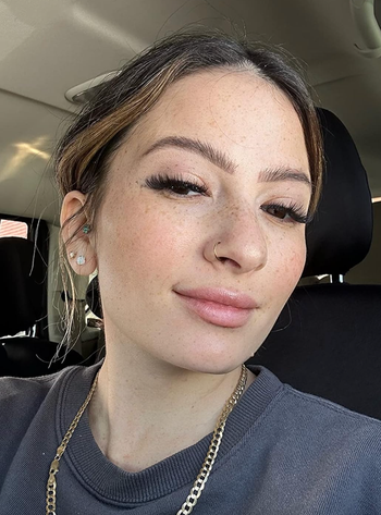 A close-up selfie of a woman showcasing her long lashes, understated makeup, a nose ring, and a gold chain necklace, dressed in a gray top with a car interior in the background.