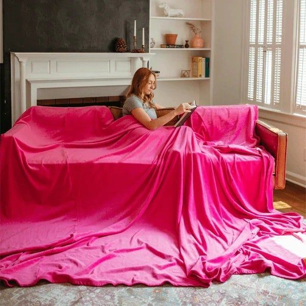 A model lounging on a sofa, reading a book while draped in an oversized pink blanket, in a cozy room setting