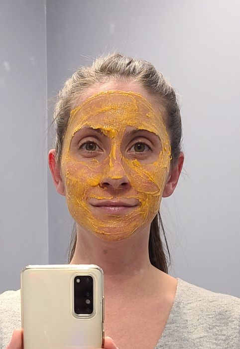 A woman with a turmeric face mask snaps a selfie in front of a mirror, her expression neutral as she holds her smartphone.