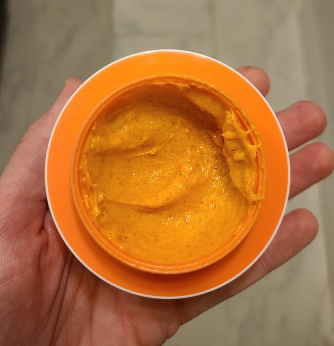 A hand is pictured with an open container of smooth, yellow-orange cream in a shopping article.