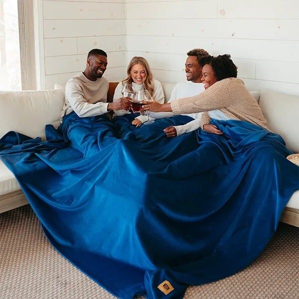 A group of four models clinking glasses under a massive blue blanket on a white sofa.