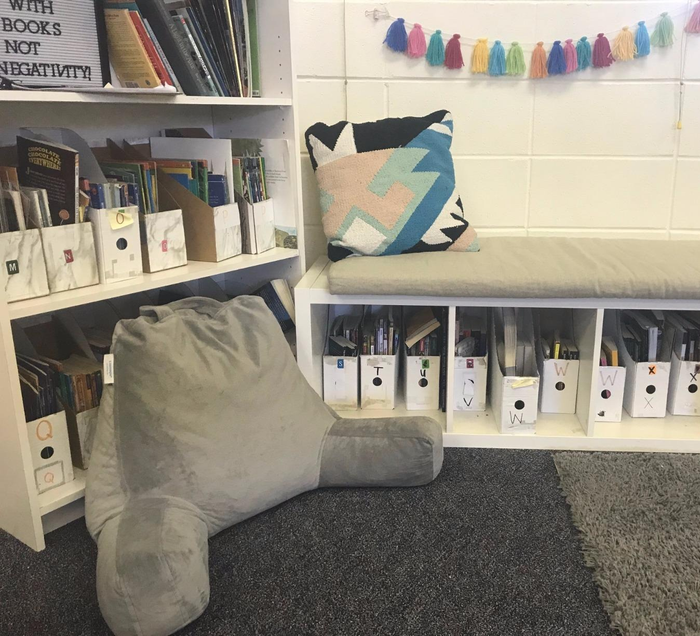 A cozy reading nook featuring bookshelves, comfy seating, and a colorful tassel garland. Boxes marked with alphabet letters hold various books.