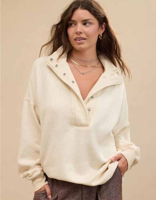 A person rocking a cozy pullover with a high collar, paired with casual trousers. They accessorize with layered necklaces and earrings, and their long, wavy hair adds to the relaxed look.