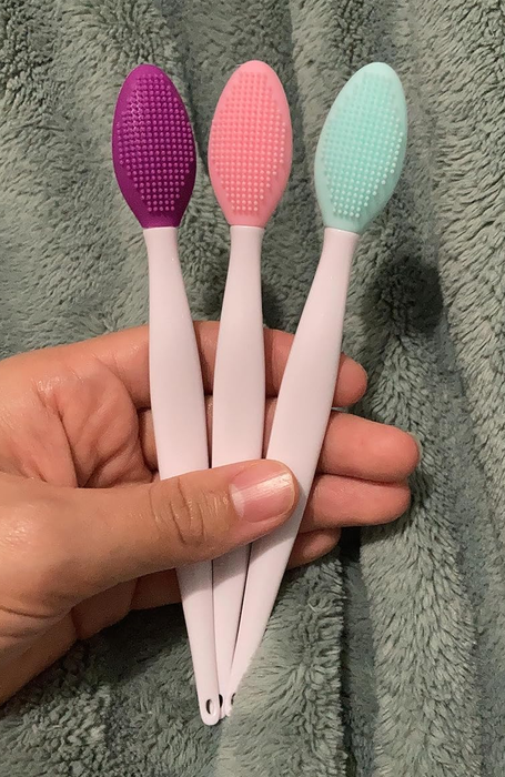 Three silicone exfoliating brushes in purple, pink, and teal, held in a hand and placed on a textured fabric background.