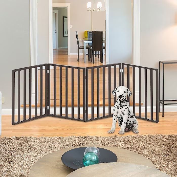 A Dalmatian puppy, gazing at the camera, sits in front of an expandable pet gate blocking a doorway