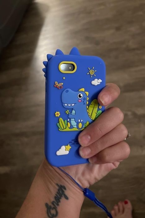 A hand gripping a phone encased in a blue dinosaur-themed cover adorned with playful dinosaurs, a sun, clouds, and butterflies. A tattoo is visible on the wrist, with the scene set indoors.