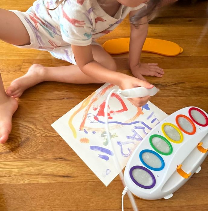 A young child sits on the floor, holding a white electronic paintbrush, creating artwork on Color Wonder paper beside a toy paint palette.