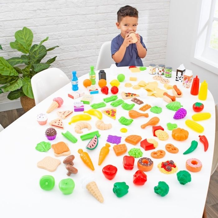 A young child sits at a table surrounded by an assortment of toy food items, such as fruits, vegetables, and snacks, immersed in imaginative play.