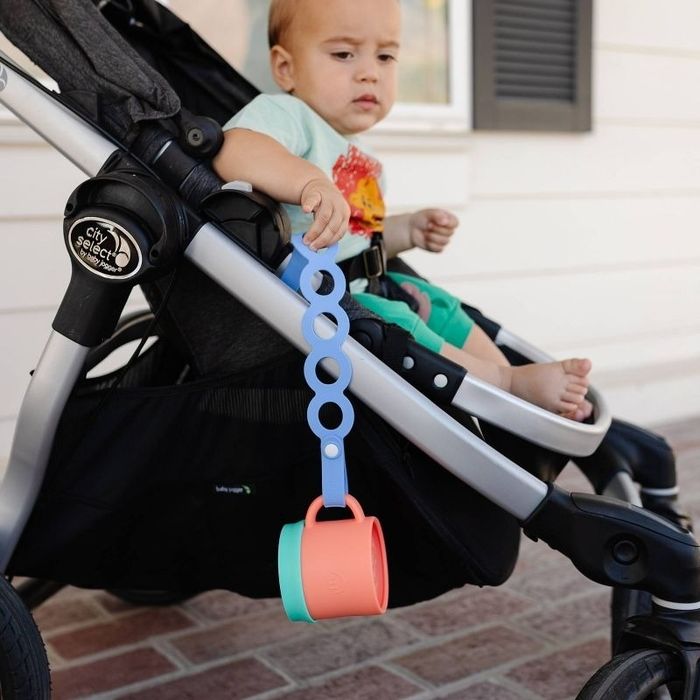 A toddler in a stroller holding a silicone teether attached to a cup strap. The article is categorized as Shopping