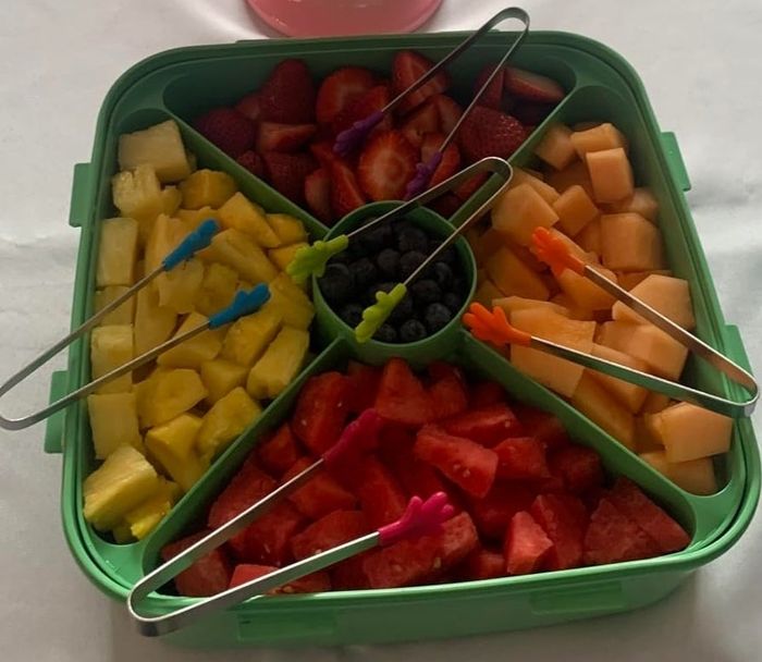 A platter filled with assorted fruits, accompanied by tongs and a cake stand topped with decorated cookies, all arranged on a tablecloth.
