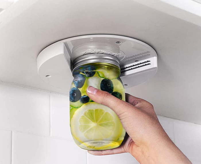 A hand using an under-cabinet jar opener to unscrew a jar filled with lemon slices and blueberries.