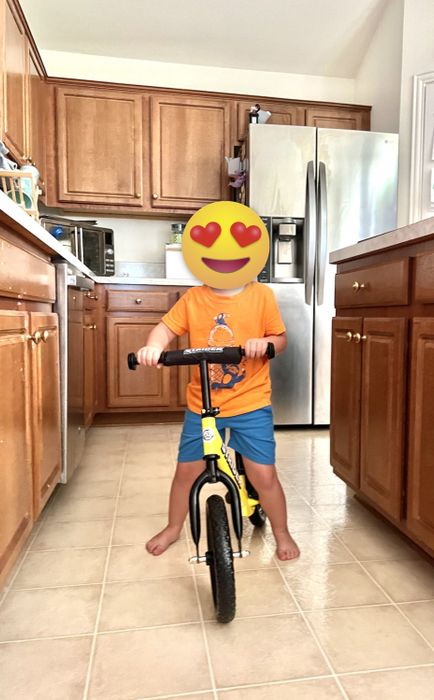 A young child rides a yellow balance bike in a kitchen, with wooden cabinets and a stainless steel refrigerator visible in the background.