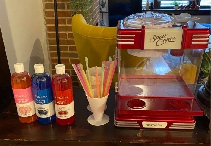 A snow cone machine paired with an assortment of syrups, including cherry, blue raspberry, and grape, along with plastic spoon straws, all beautifully displayed on a kitchen counter in a homey setting.