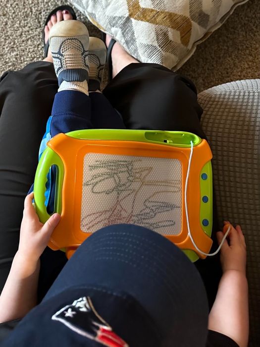 A child wearing a Patriots cap and socks draws on a magnetic sketch pad while sitting on an adult’s lap.