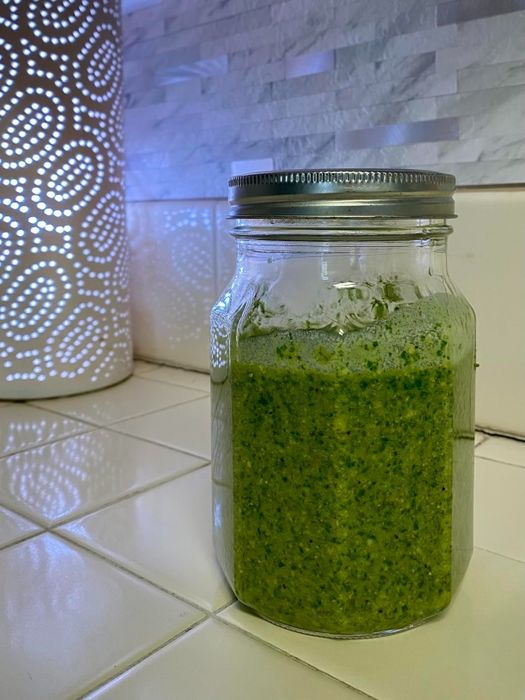 A glass jar of vibrant green pesto sauce rests on a kitchen counter, beside a beautifully designed lamp, creating a warm, inviting atmosphere.