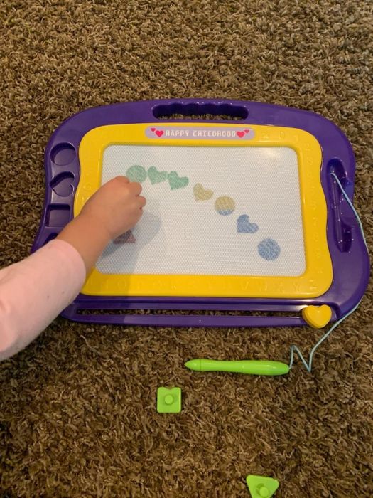A child’s hand uses a green stylus to draw shapes and hearts on a purple and yellow magnetic drawing board. The top of the board displays the text 