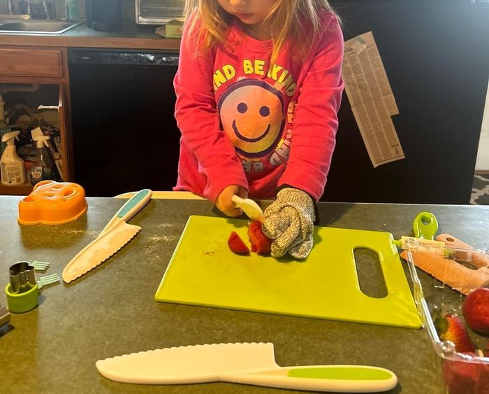 A young child in a kitchen, carefully slicing a strawberry on a green cutting board while wearing a protective glove. A pumpkin-shaped item and knife rest on the counter.