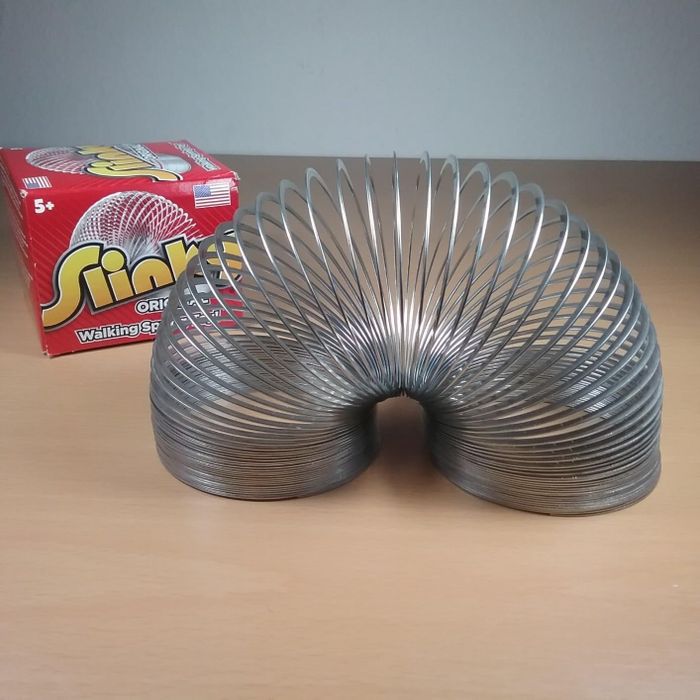 A Slinky toy positioned in the foreground with its red and yellow packaging visible in the background, placed on a wooden surface. This is part of an article discussing classic toy shopping.