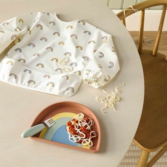 A children's rainbow-printed long-sleeve bib sits on a table next to a used plate of spaghetti with sauce, some noodles scattered across the table