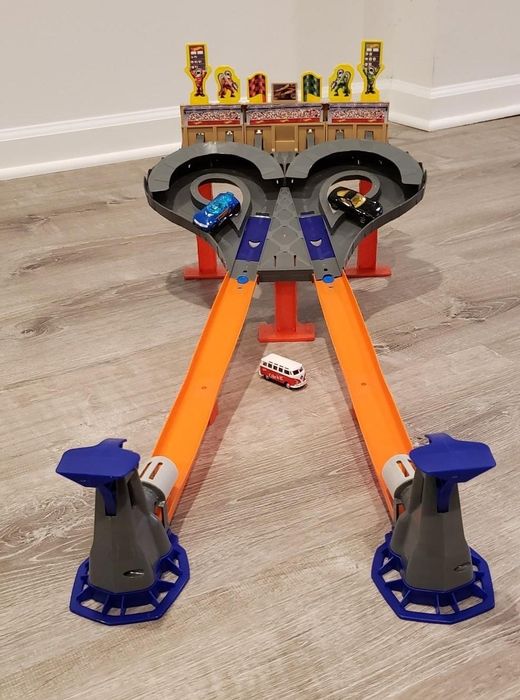 A toy car racing set featuring two vibrant orange lanes and blue launchers, with toy cars ready on the track and a few scattered on the floor beneath.