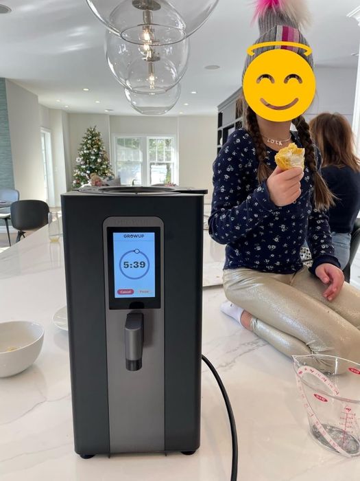 A child (with their face hidden) sits on a kitchen counter, enjoying an ice cream cone beside a Grown-Up soft serve machine, with a Christmas tree in the background.