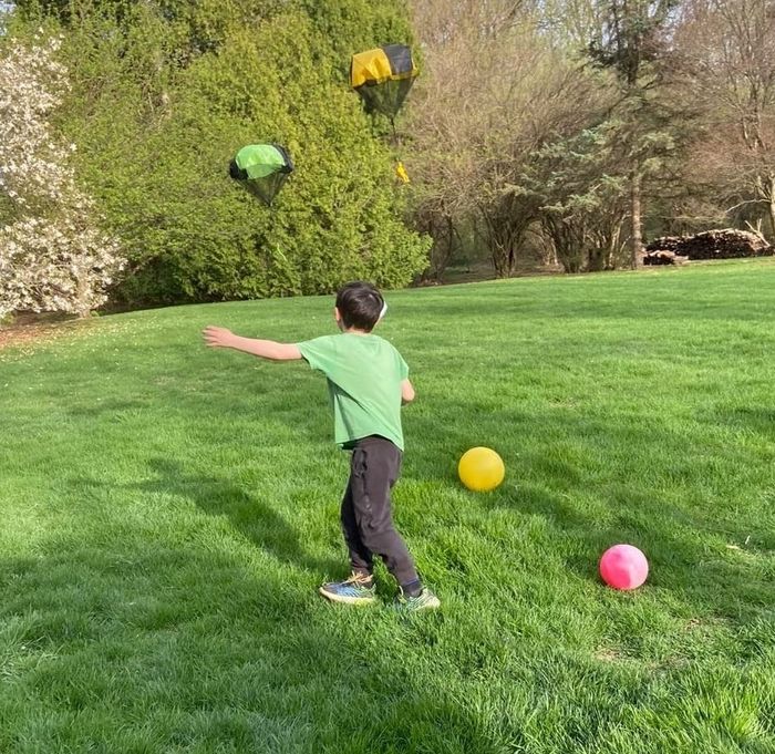 A child wearing a green shirt plays on a lawn, launching colorful parachute toys into the air, surrounded by yellow and pink balls.