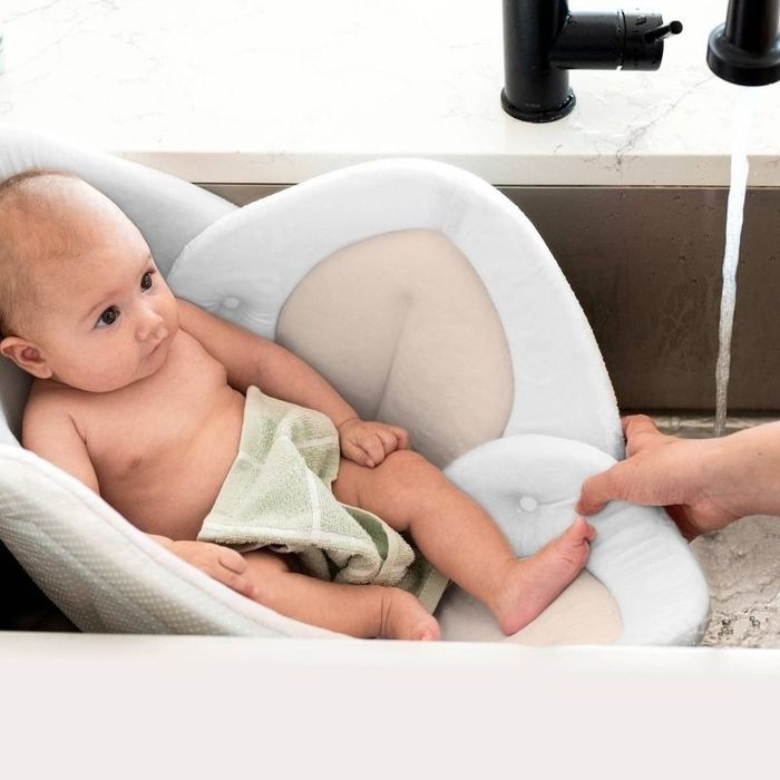 A baby sits comfortably in a soft, padded bath seat inside a sink, as an adult adjusts the temperature of the water. A green washcloth is draped over part of the baby’s torso.