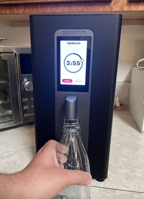 A person holds a glass bottle under a 'Growup' water dispenser, which displays a countdown timer set to 3:55, with a microwave visible in the background.