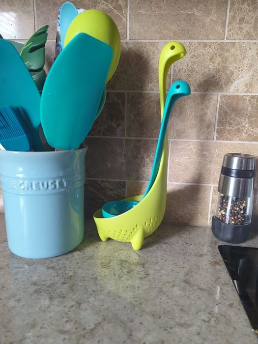 A Loch Ness monster-themed kitchen ladle in vibrant lime green, placed beside a blue utensil holder filled with various cooking tools and a pepper grinder on the counter.