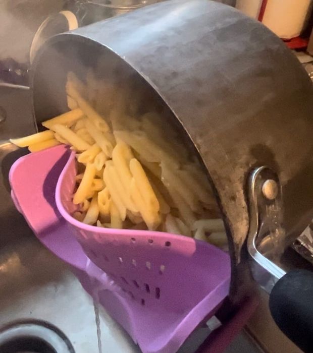 A pot of penne pasta is being drained into a purple colander over the sink in the kitchen.