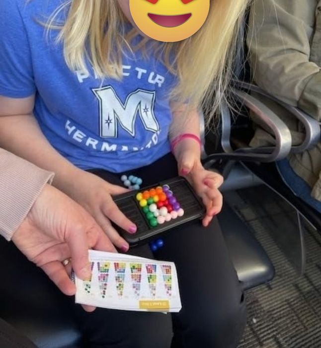 A young child holds a colorful bead puzzle toy, while an adult hand displays a card with bead arrangements. The child is intently focused on solving the puzzle.