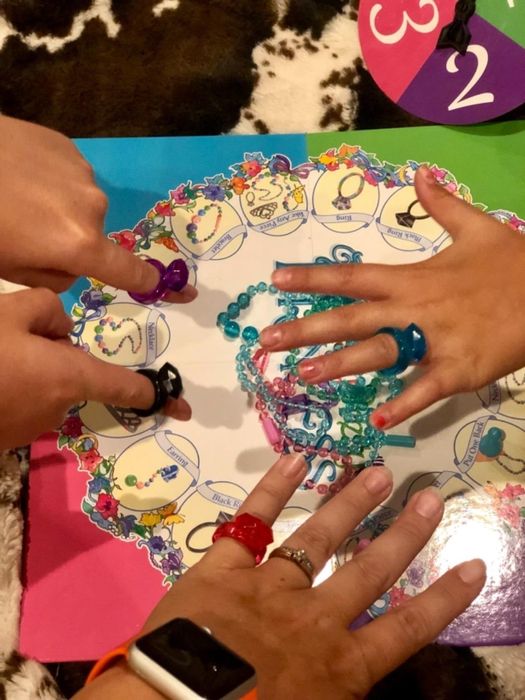Multiple hands adorned with rings engage in a bracelet-making board game.