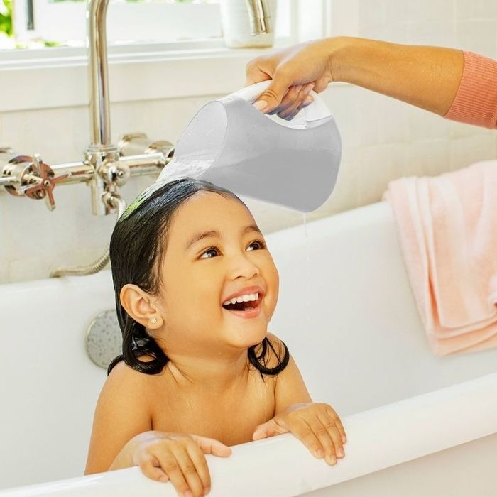 A young child grins happily while being bathed in a bathtub, as an adult gently pours water over their head from a pitcher.