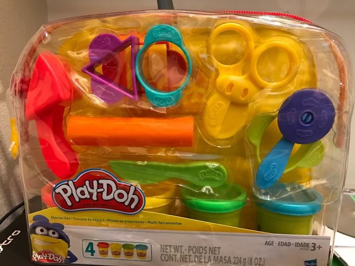 A Play-Doh set packaged in a clear plastic bag, complete with shape cutters, a roller, scissors, and small containers of Play-Doh. Designed for kids aged 3 and above.