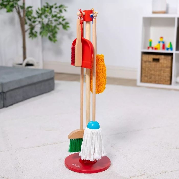 A toy cleaning set featuring a broom, mop, dustpan, and duster, all standing upright on a red base. Perfect for children’s pretend play in a living room environment.