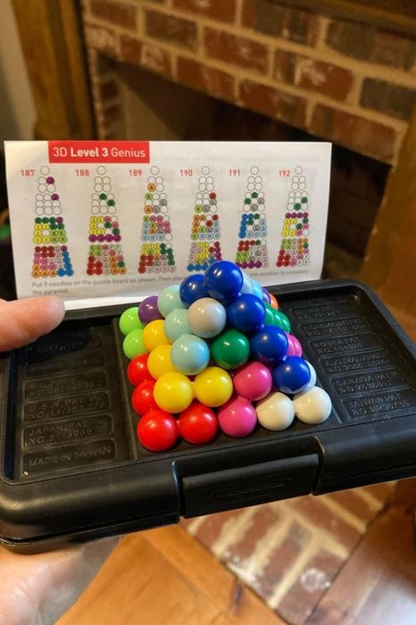 A hand gripping a sleek black puzzle case filled with vibrant, stacked marbles, alongside a 3D Level 3 Genius challenge sheet displaying various intricate configurations.