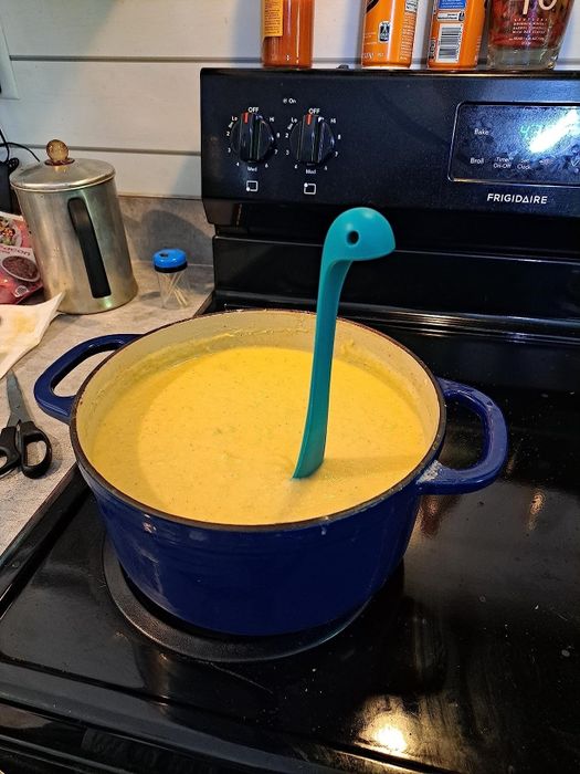A blue pot filled with smooth, creamy soup sits on the stove, with a blue dinosaur-shaped ladle resting inside. The background shows various kitchen appliances and utensils.