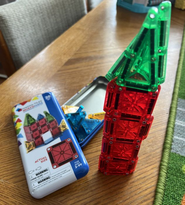 A magnetic construction set from Magna-Tiles, featuring a partially built 3D structure, placed on a wooden table with an open tin box revealing additional pieces.