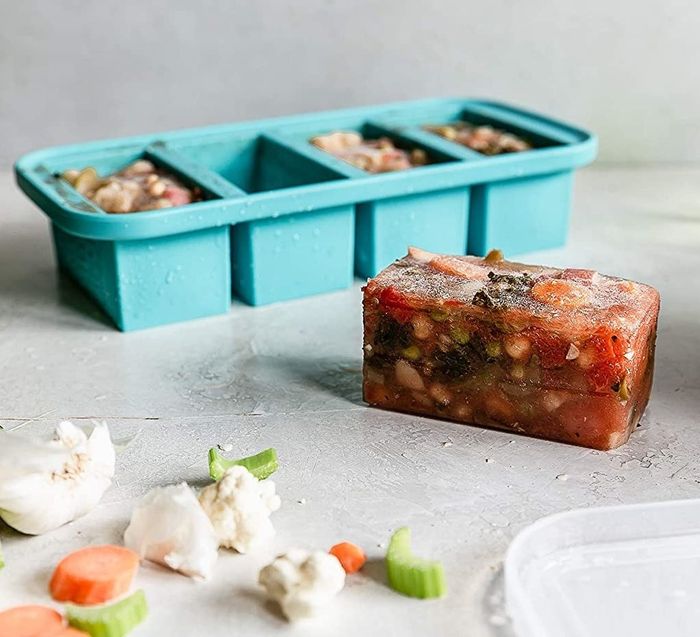 A five-compartment teal tray resembling a large ice cube tray, with a block of frozen soup neatly placed beside it.