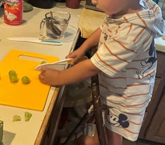 A young child stands on a chair in the kitchen, cutting Brussels sprouts on a yellow cutting board, wearing a shirt with a Mickey Mouse design.