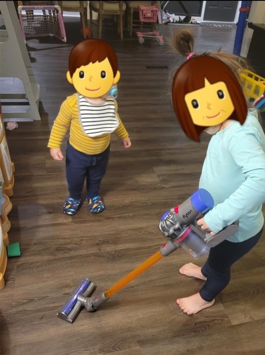 Two kids play with a toy vacuum cleaner in a living room. The older child is vacuuming while the younger one observes curiously.