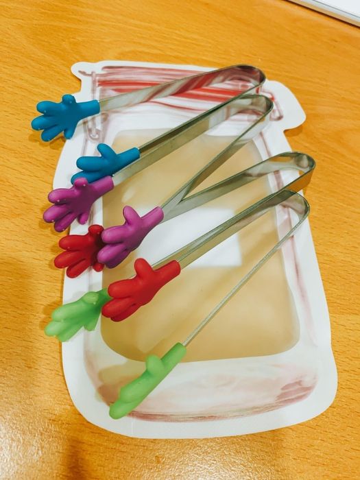 Brightly colored silicone tongs shaped like hands, with metal handles, placed on a table beside a jar illustration.