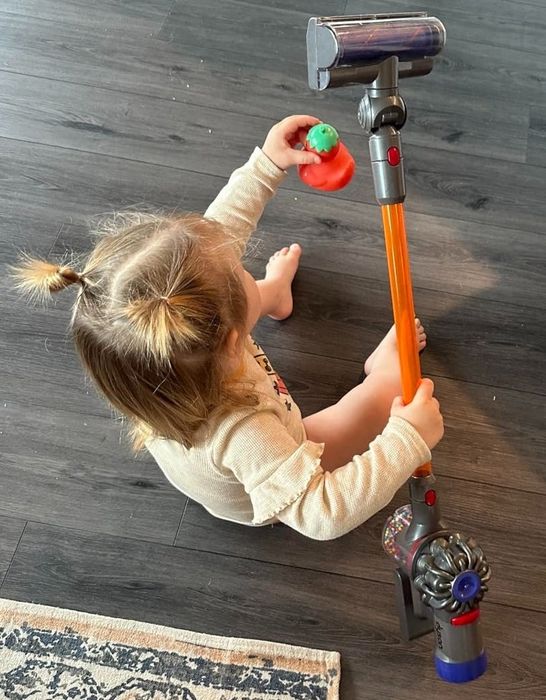 A pigtailed child sits on the floor, holding a small vacuum cleaner and a toy.