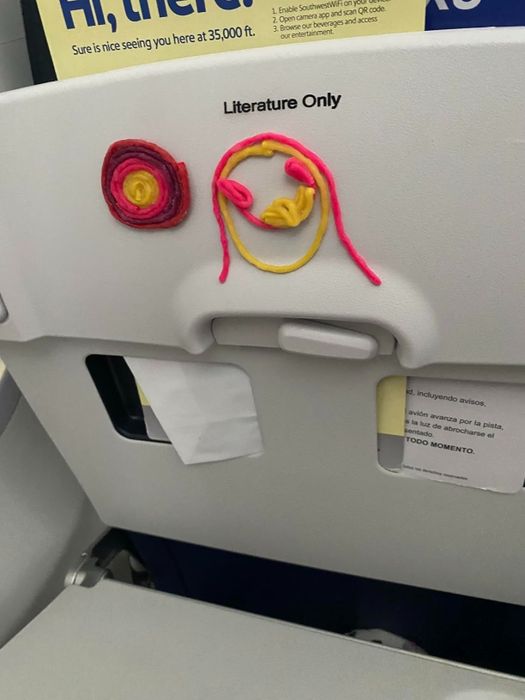 An artistic creation using Wikki Stix, showcasing the abstract faces of a woman and child, is affixed to the back of an airplane seat, just above a pocket marked 