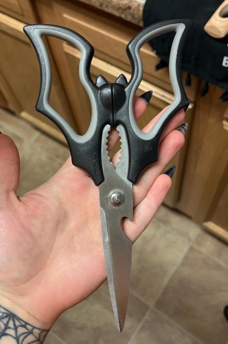 A hand holds a pair of sleek black kitchen shears with a built-in bottle opener, positioned next to a kitchen sink for easy access.