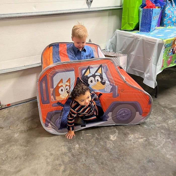 Two children play inside a Bluey-themed car tent, surrounded by gift bags and a decorated table in the background.