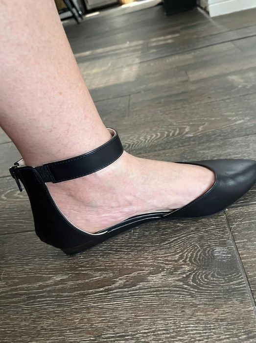 Close-up of a foot adorned with a black leather flat shoe featuring an ankle strap, set on a wooden floor.