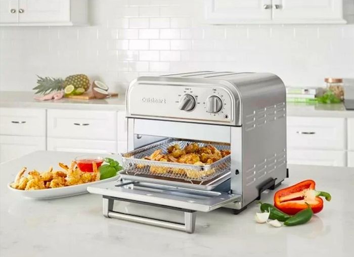 A shot of an air fryer in use with crispy fried food inside, accompanied by vegetables and a plate of food next to it